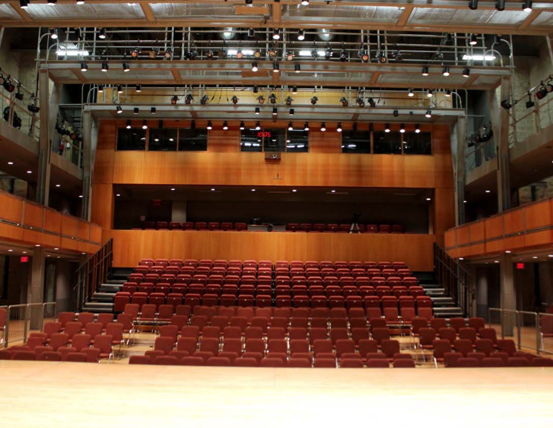 View from the stage looking out at an empty auditorium with rows of red seats and overhead lighting rigs.